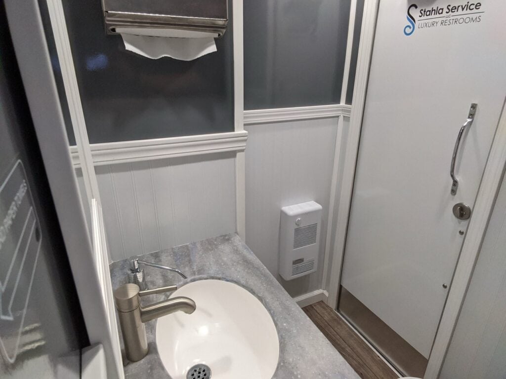 a restroom with a white sink, countertop, paper towel dispenser, and a door labeled "stahla service luxury restrooms." the sink has a stainless steel faucet, and there's a heater mounted on the wall.