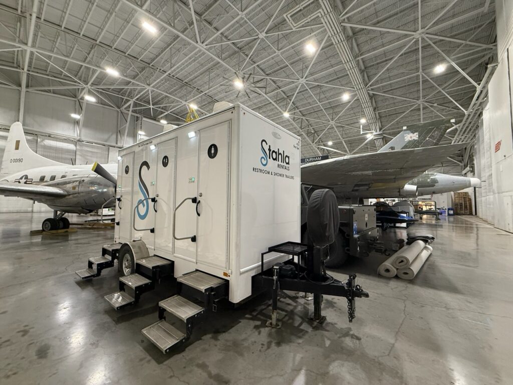 A portable restroom trailer parked inside an aircraft hangar next to large airplanes, with supplies and equipment visible nearby.