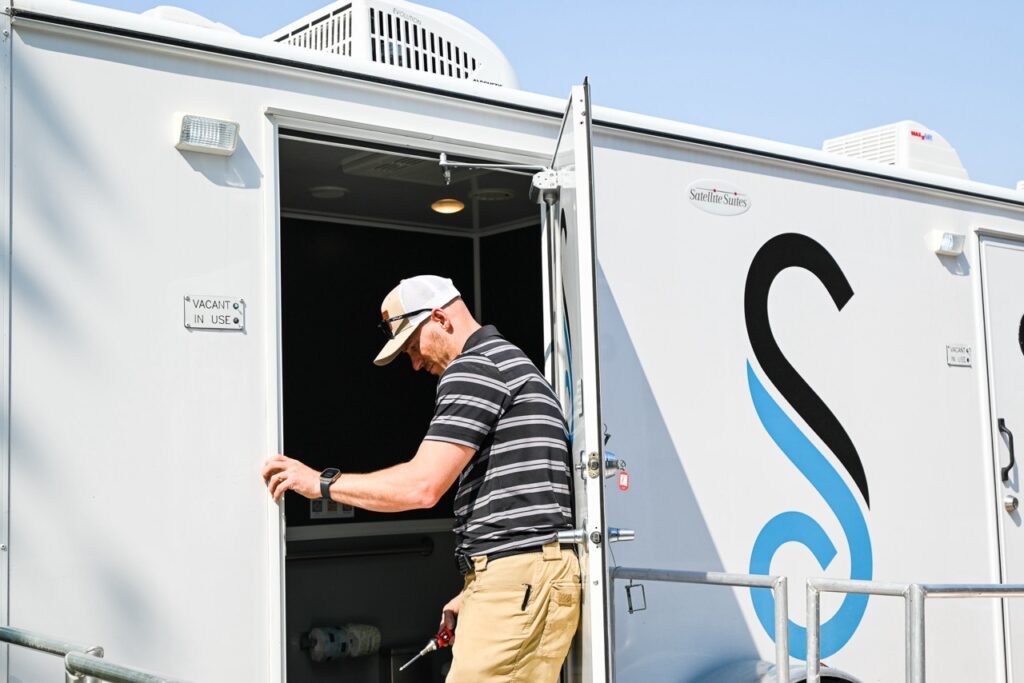 a man in a striped shirt and baseball cap stands near the open door of a portable restroom unit, provided by stahla services, at the orange city tulip festival, blending comfort and tradition effortlessly.