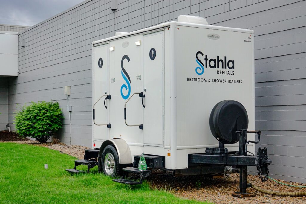a portable restroom and shower trailer, branded "stahla services," is parked beside a grey building in omaha and connected to a hose.