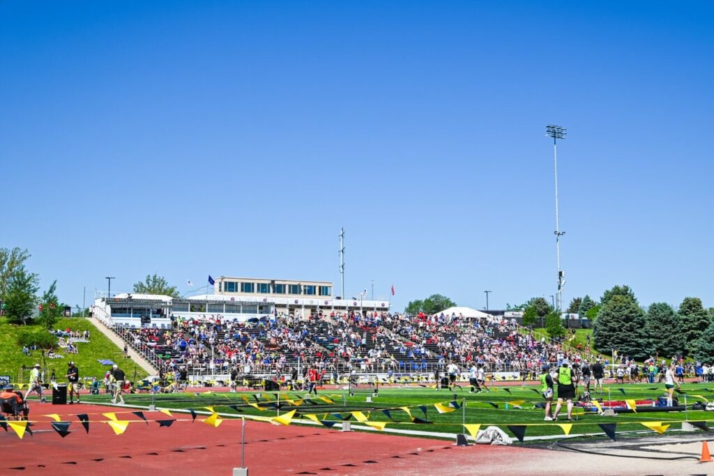a large crowd sits in bleachers at an outdoor sports stadium on a sunny day for the nsaa state track event, with people on the field and surrounding areas, and triangular flags hanging along the track. the attendee experience is enhanced by convenient restroom trailers nearby.