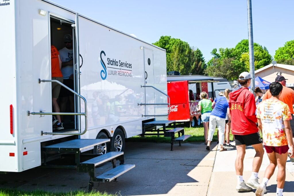 people standing and walking around a parked mobile unit with "shahla services luxury restrooms" and a red beverage truck at the nsaa state track event 2024, enhancing the attendee experience at this outdoor event.