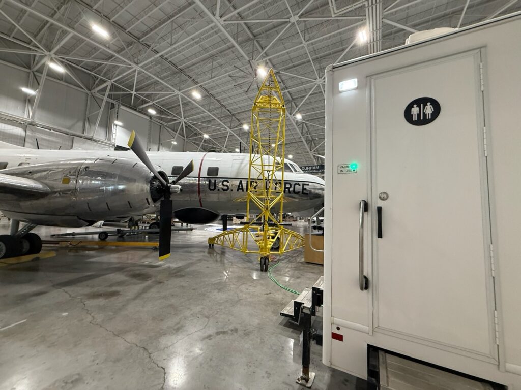 A vintage u.s. air force aircraft is displayed in a hangar next to a portable restroom trailer and yellow maintenance scaffolding.