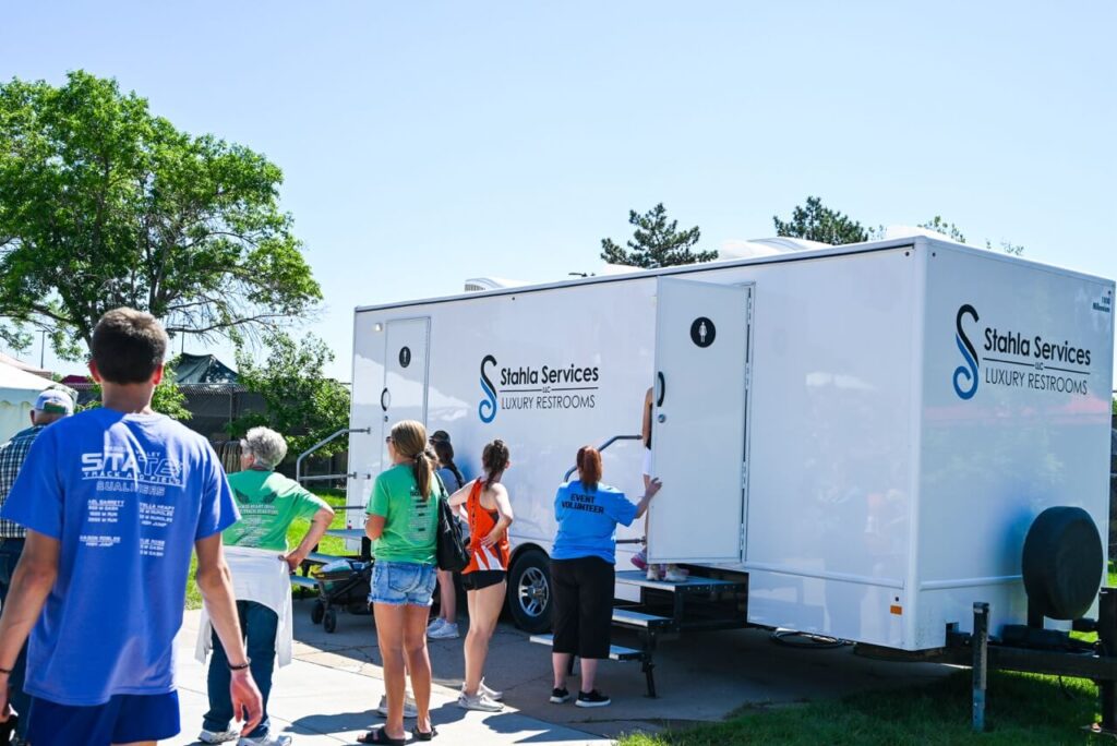attendees at the nsaa state track event stand in line in front of a mobile restroom trailer operated by stahla services, enhancing the overall attendee experience on a sunny day with trees in the background.