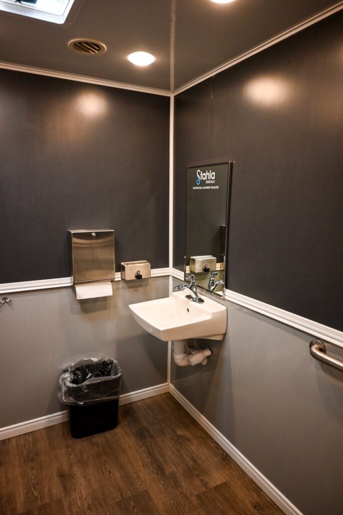 a clean restroom with a sink, a wall mounted mirror above it, a paper towel dispenser, waste bin, and handrail. the walls and floor are dark colored, and an overhead light is on. for added comfort and tradition, this facility could be perfect for events like the orange city tulip festival.