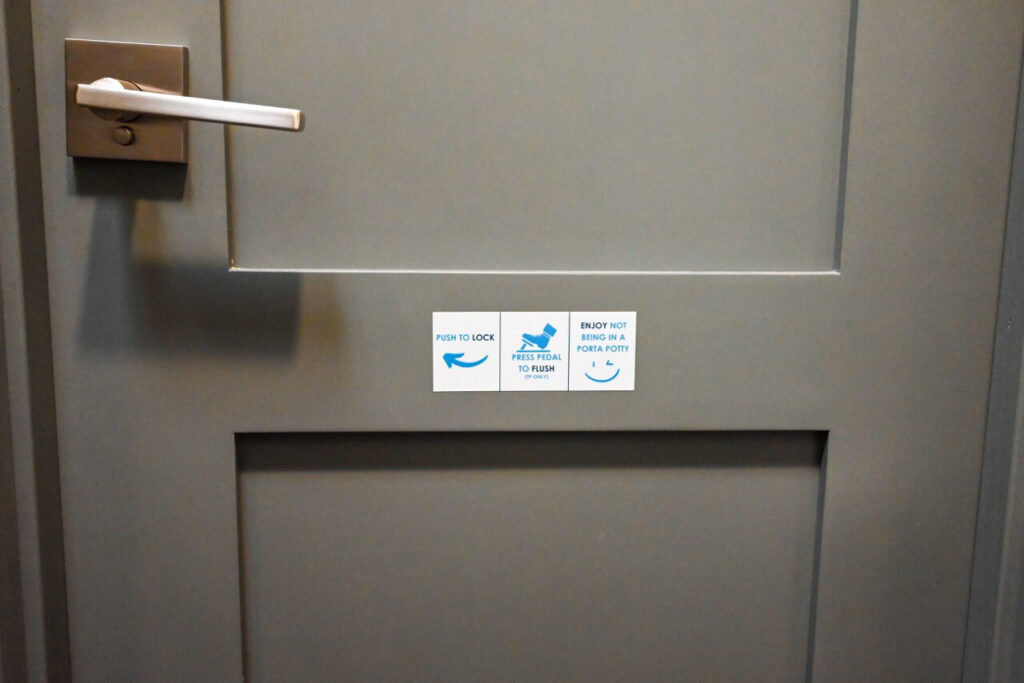 gray bathroom door with three instructional stickers: "push to lock," "press pedal to flush," and "enjoy not being in a porta potty" accompanied by respective icons. perfect for enhancing attendee experience at events like the nsaa state track event 2024 with clean restroom trailers.