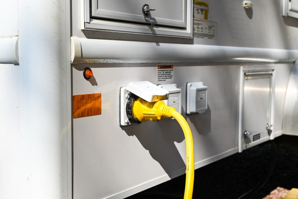 close up of an electrical connection with a yellow power cord plugged into an rv, highlighting the comfort and tradition of road trips. various sockets and labels are visible on the side of the vehicle.