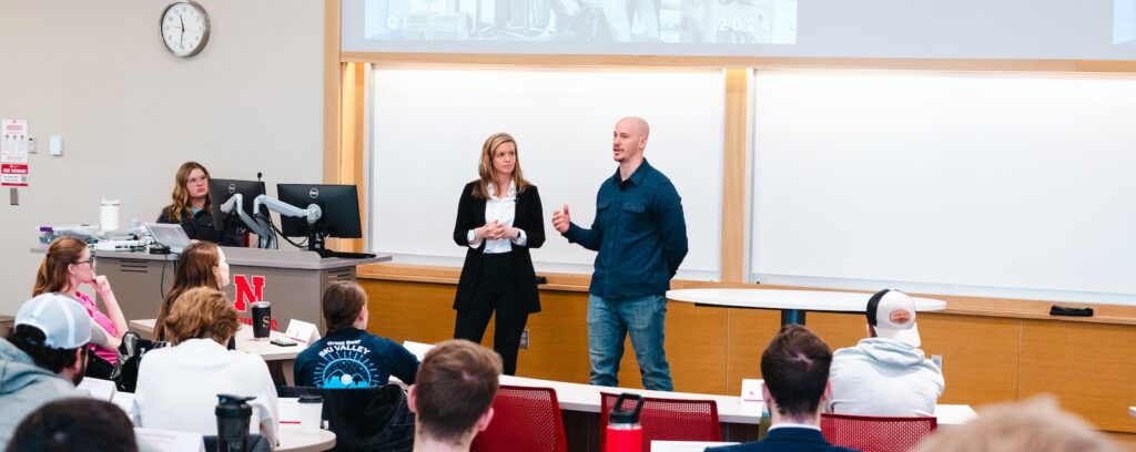 Two People Are Speaking In Front Of A Classroom. A Woman Stands To The Left, And A Man Is Gesturing To The Right. Several Students Are Seated, Facing Them.