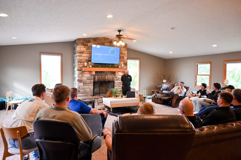 A group of people sit in a living room facing a presenter who stands near a fireplace and a TV screen displaying a presentation.