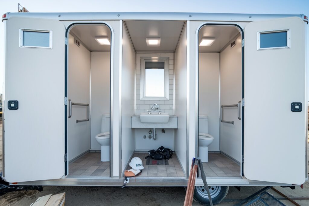 image of a portable restroom trailer often found on construction sites, featuring three compartments—two with toilets and one with a sink. a hard hat and backpack are placed on the floor in front of the sink, highlighting it's importance for workers without hidden costs.