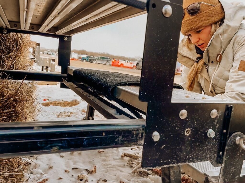 A person in winter clothing works on or inspects a metal structure outdoors in a snowy area with hay bales and parked cars in the background.