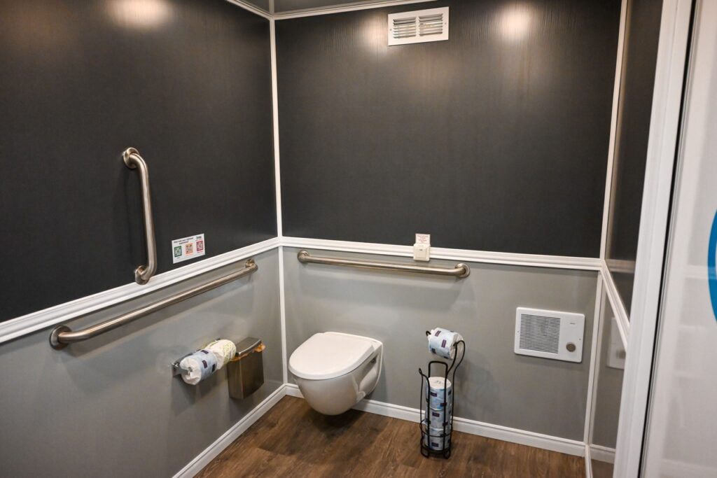 a modern bathroom with wall mounted toilet, handrails, toilet paper holder, and a trash bin. the floor is wooden, and the walls are dark with white trim. this space effortlessly blends comfort and tradition, reminiscent of the warm hospitality found during the orange city tulip festival.