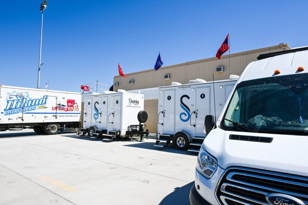 several parked trucks and restroom trailers with different company logos and flags on top next to a beige building under a clear blue sky enhanced the nsaa state track event attendee experience.