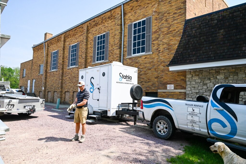 a man in a striped shirt and cap stands beside a white trailer and pickup truck outside a brick building with large windows, embodying the comfort and tradition of the area.