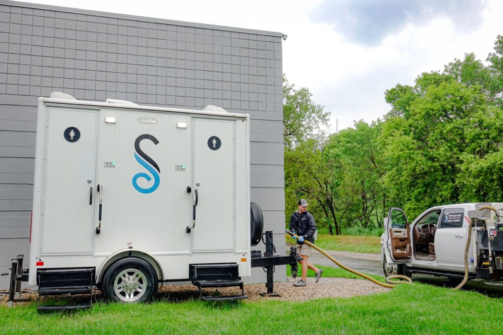 a person connects a hose to a white portable restroom trailer next to a truck in an outdoor setting with grass, trees, and an overcast sky, likely provided by stahla services.