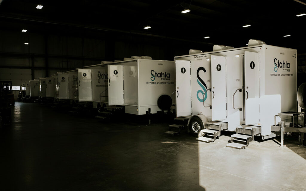 Several white portable restroom and shower trailers are lined up inside a large, dimly lit warehouse. Sunlight partially illuminates the row of trailers on the right side.