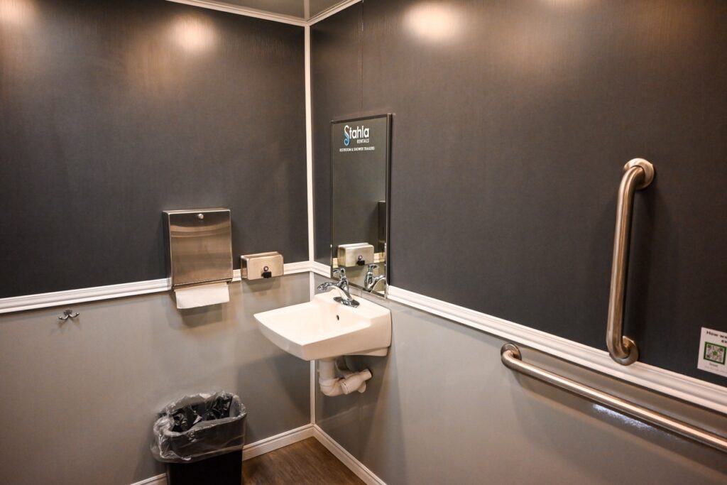 a small bathroom with dark gray walls featuring a wall mounted sink, a mirror, a paper towel dispenser, a trash bin, a grab bar, and a soap dispenser—a seamless blend of comfort and tradition reminiscent of the meticulous standards maintained by stahla services.