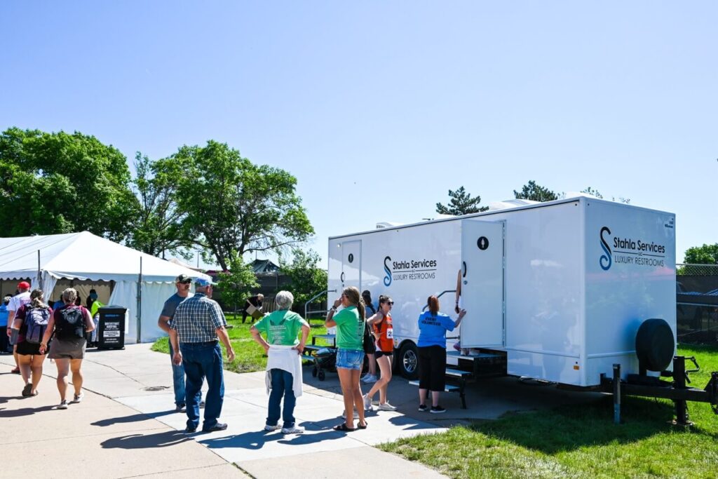 people are standing in line outside a mobile restroom unit labeled "stahla services luxury restrooms" at the nsaa state track event, enhancing the attendee experience amidst a backdrop of white tents and trees.
