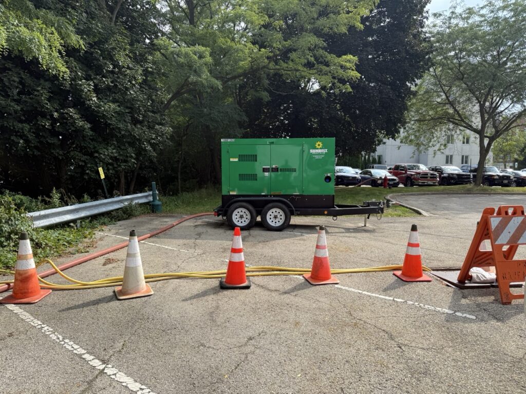 A green pull behind generator on a trailer is parked in a lot, surrounded by orange traffic cones and caution tape, with cables running on the ground—ideal for powering shower trailers or restroom trailers at event sites.