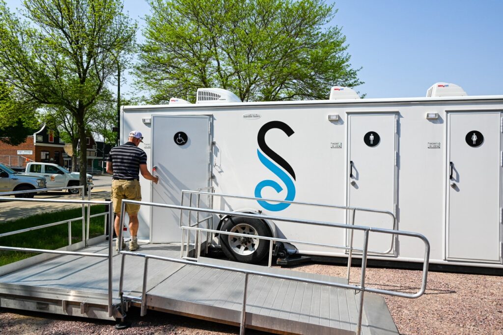 a man in a striped shirt and shorts stands at the entrance of an accessible portable restroom unit with a wheelchair ramp outside, provided by stahla services, ensuring comfort and tradition are maintained at the orange city tulip festival.