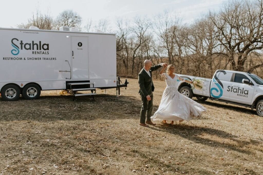 A bride and groom joyfully dance outside near luxury portable toilets for weddings in a barren landscape.