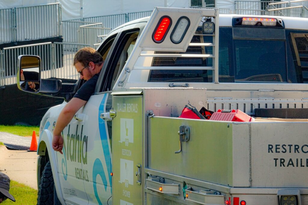 A man in glasses sits in a pickup truck with company branding, reaching out the window, while equipment for staff training is stored in the truck bed’s side compartment.