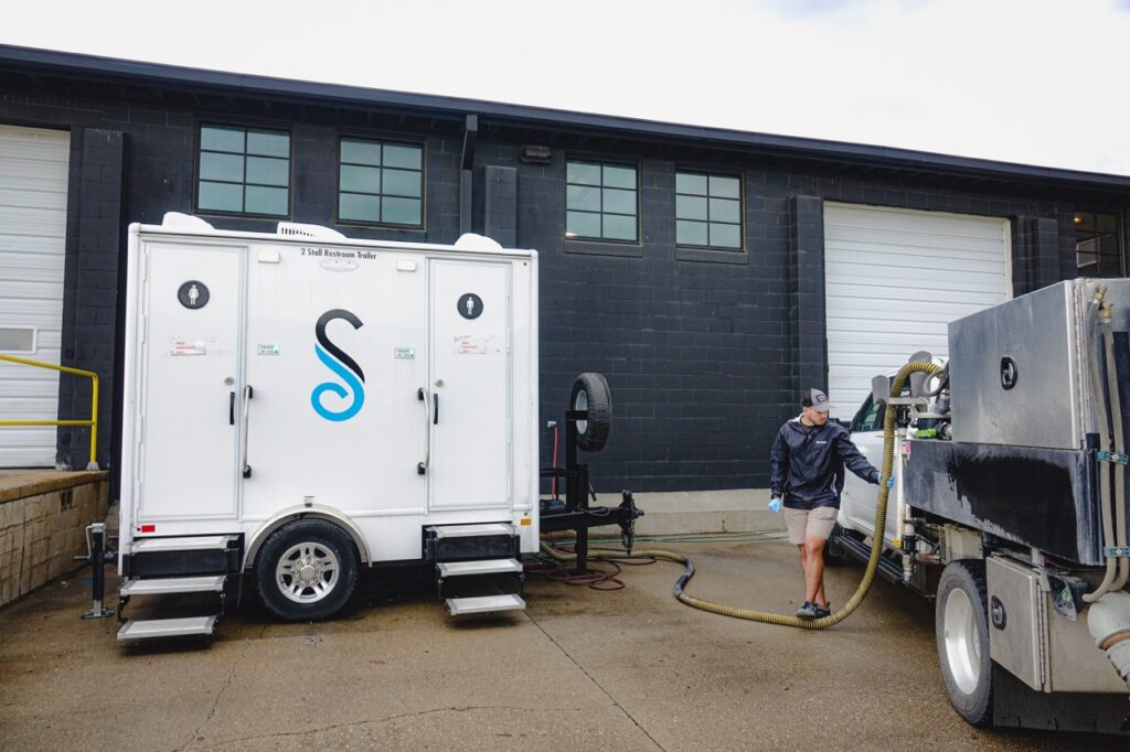 Image 8: a worker from stahla services meticulously services a mobile sanitation unit stationed outside a red bull distribution company building with large garage doors in omaha. the worker is handling a hose connected to their nearby truck, ensuring everything runs smoothly.