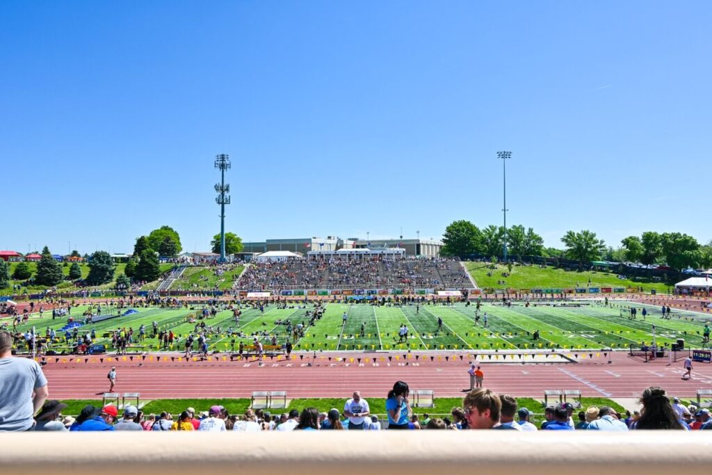 a wide shot of the nsaa state track event on a clear day. the sports stadium is filled with spectators, and several athletes are preparing on the field. restroom trailers are strategically placed to enhance the attendee experience.