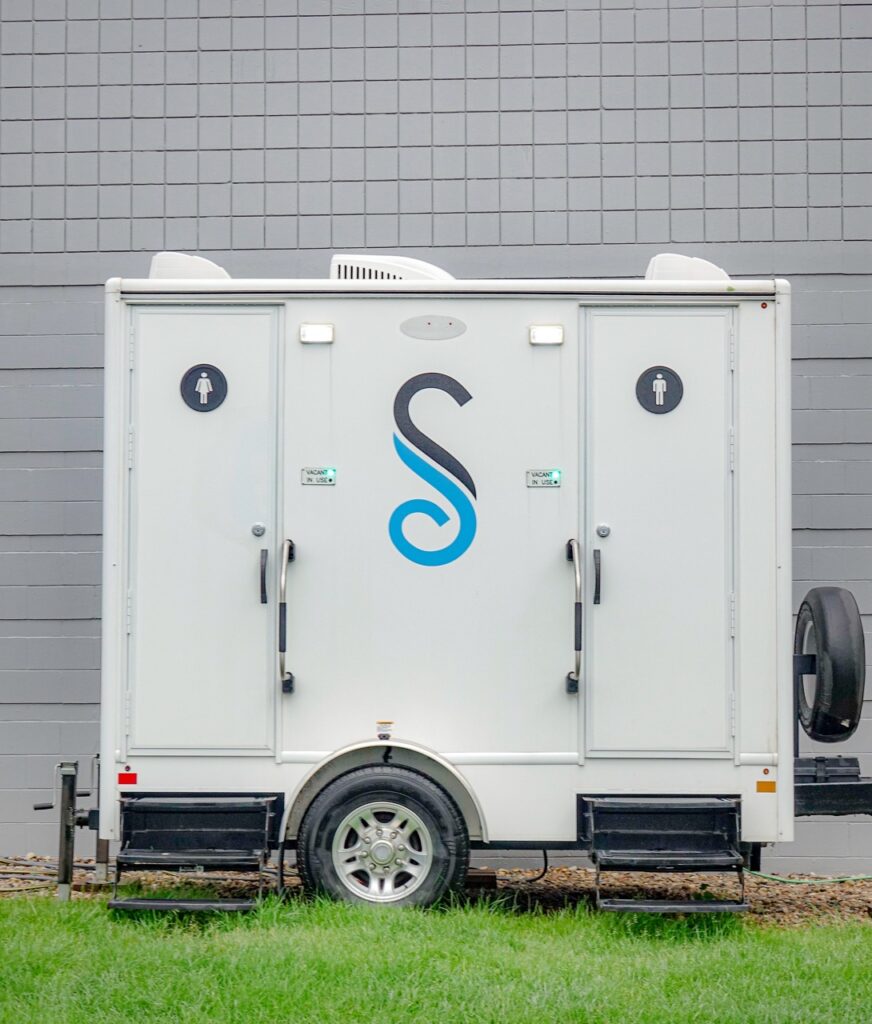a mobile restroom trailer from stahla services with separate entrances for men and women stands against a gray tiled wall. the unit, conveniently placed near h&h kia omaha, has an "s" logo and a small set of steps leading up to each door.