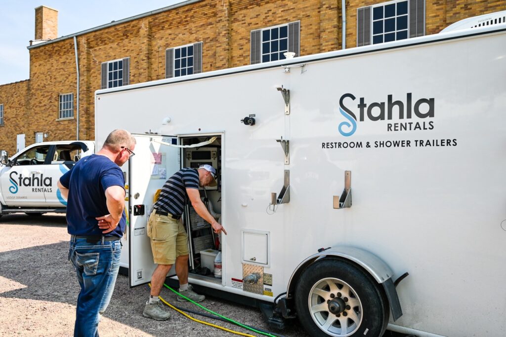 two men inspect equipment stored in a white stahla rentals trailer, marked for restroom and shower use, parked in front of a brick building. another stahla rentals vehicle is visible in the background, showcasing the comfort and tradition that stahla services brings to events like the orange city tulip festival.