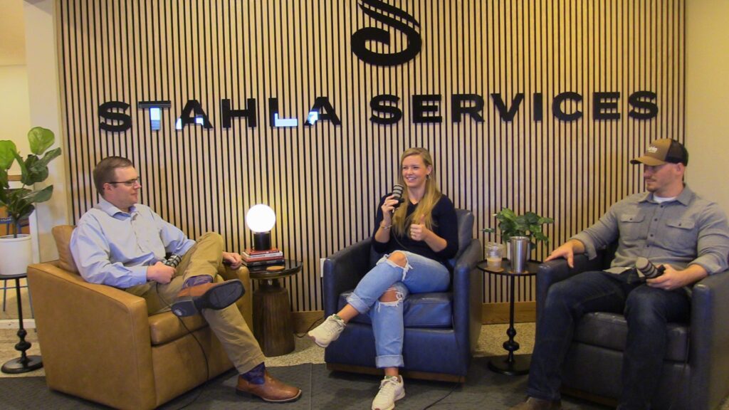 Three people are seated in armchairs, having a conversation with microphones in front of a wall that reads "Stahla Services," discussing Building Nebraska through excellence and growth.