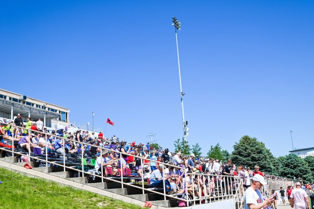 a large crowd of people seated on stadium bleachers under a clear blue sky at the nsaa state track event 2024. some attendees are standing and talking, while others are seated and watching the event unfold. tall lights and trees are in the background, enhancing the attendee experience.