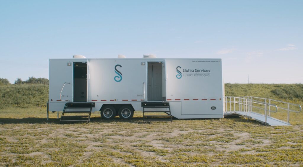 a white 10 stall restroom trailer with steps and a ramp, labeled "stahla services luxury restrooms," is parked on a grassy field under a clear blue sky.