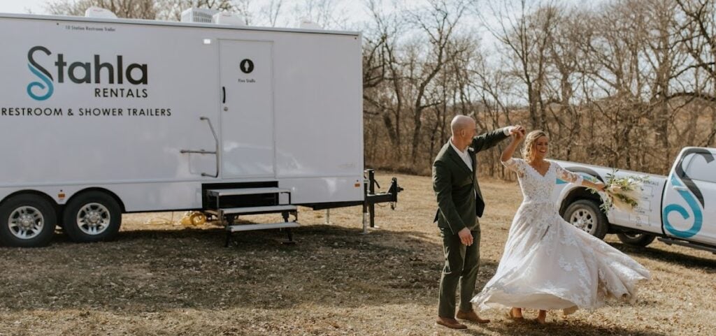 Bride and groom dancing near rental restroom trailer outdoors.