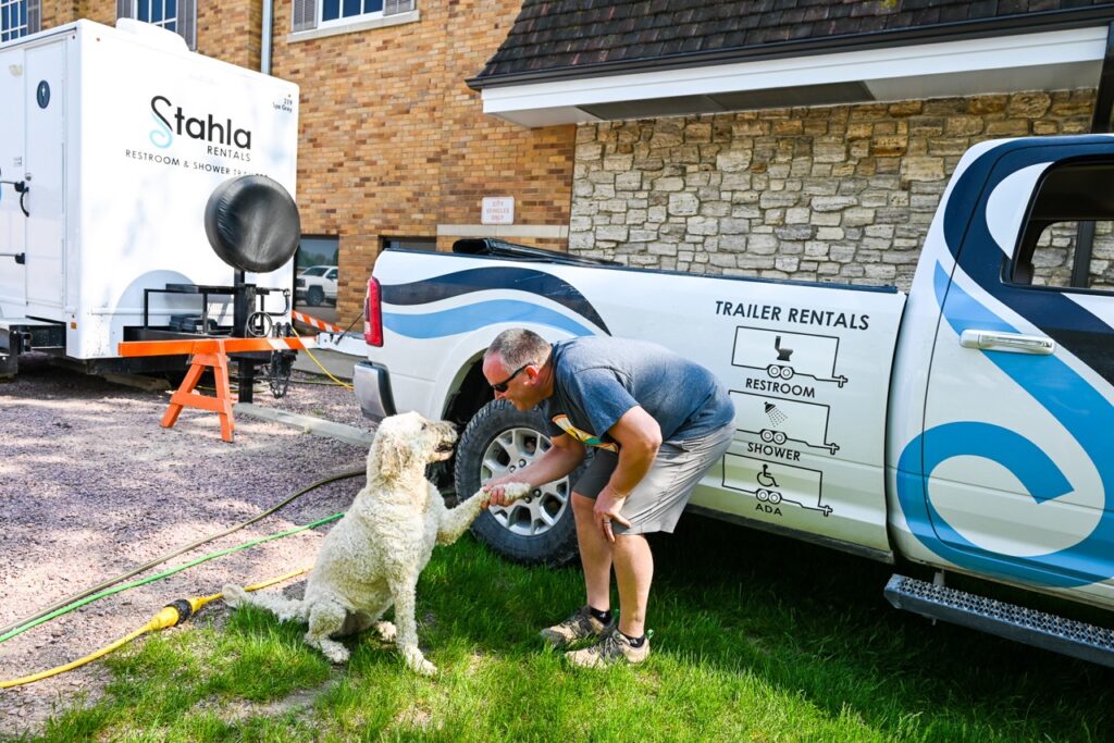 a man shakes hands with a white dog beside a trailer rental truck parked on grass, with a restroom trailer by stahla services and a building in the background, reflecting the comfort and tradition found at the orange city tulip festival.