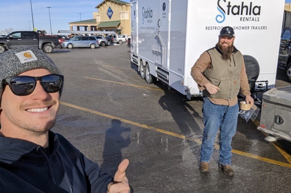 Two men posing near a rental trailer in parking lot.