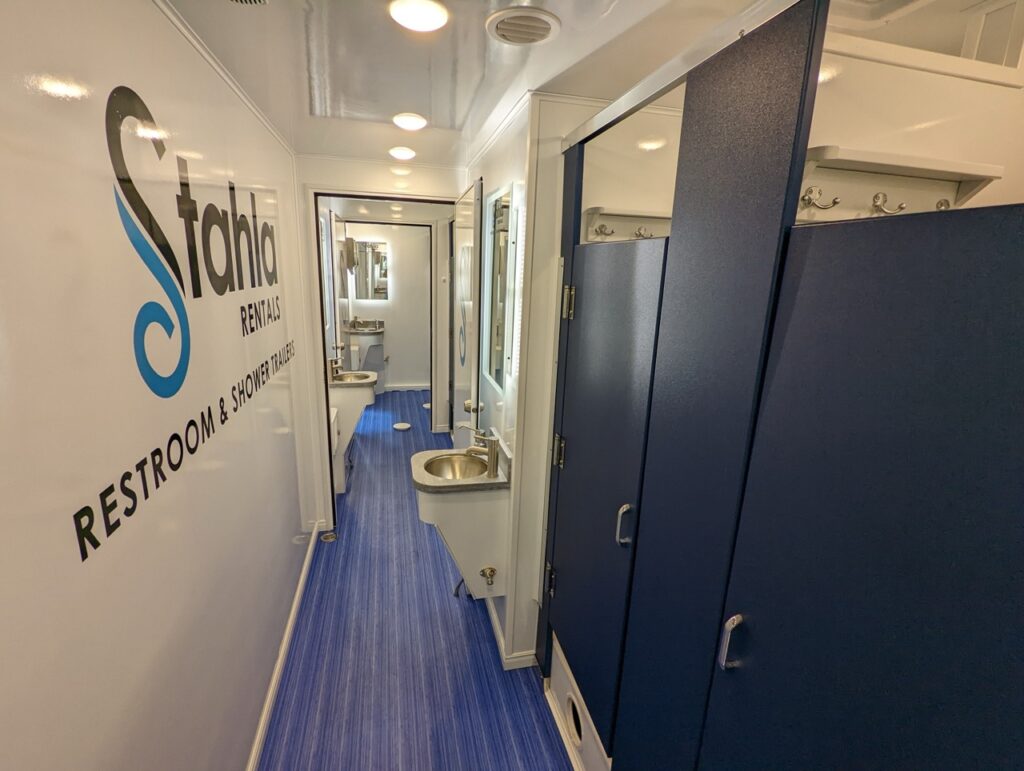 interior view of a mobile shower unit with blue flooring, stalls, and a sink. the wall features the logo and name of the stahla services, showcasing their expertise in providing top tier mobile shower units.