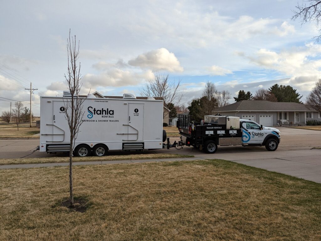 a truck with "stahla rentals" branding is towing a large, white trailer labeled "bathroom & shower trailers," indicating it's an 8 stall restroom trailer rental, parked on the side of a suburban street with houses and trees in the background.