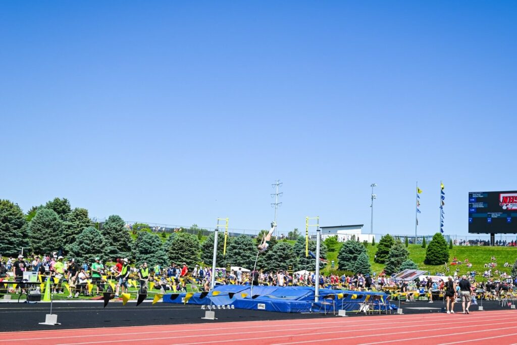 athletes compete in a high jump event at an outdoor track and field stadium during the nsaa state track event 2024, with spectators enjoying the attendee experience and a scoreboard visible in the background under a clear blue sky.