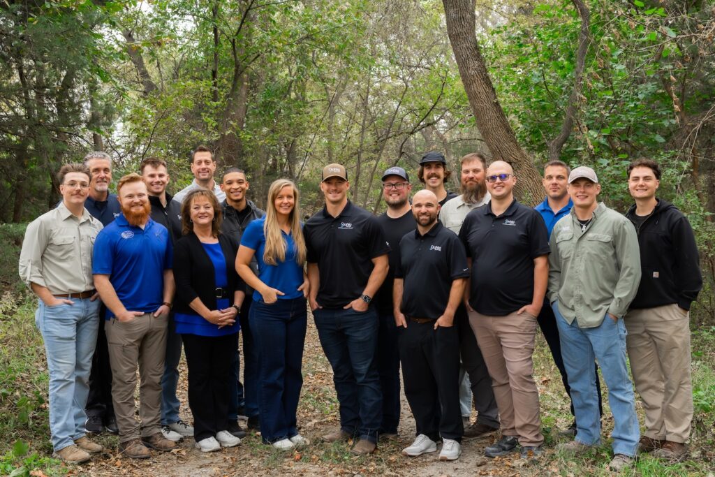 A group of seventeen adults stands together outdoors on a wooded path, smiling at the camera. Most are wearing casual shirts in blue, black, or neutral tones.
