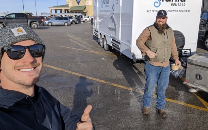 Two men smiling near trailer in sunny parking lot.