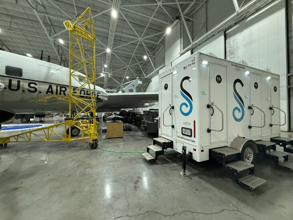 A mobile restroom trailer is parked inside an aircraft hangar next to a u.s. air force plane and yellow scaffolding.