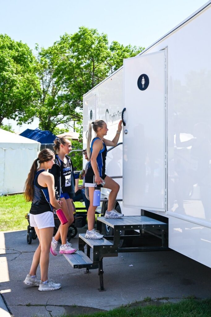 three female athletes wait in line to enter a portable restroom trailer, provided by stahla services, at the nsaa state track event. the well maintained facilities ensure an improved attendee experience for everyone present.