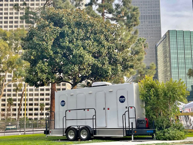 A large mobile restroom trailer is parked on grass near trees and tall office buildings in an urban setting.
