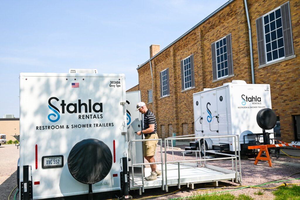 two restroom and shower trailers from stahla services are parked outdoors. a person wearing a striped shirt and shorts is standing on a ramp leading to one of the trailers. these units, perfect for events like the orange city tulip festival, offer comfort and tradition in every detail.