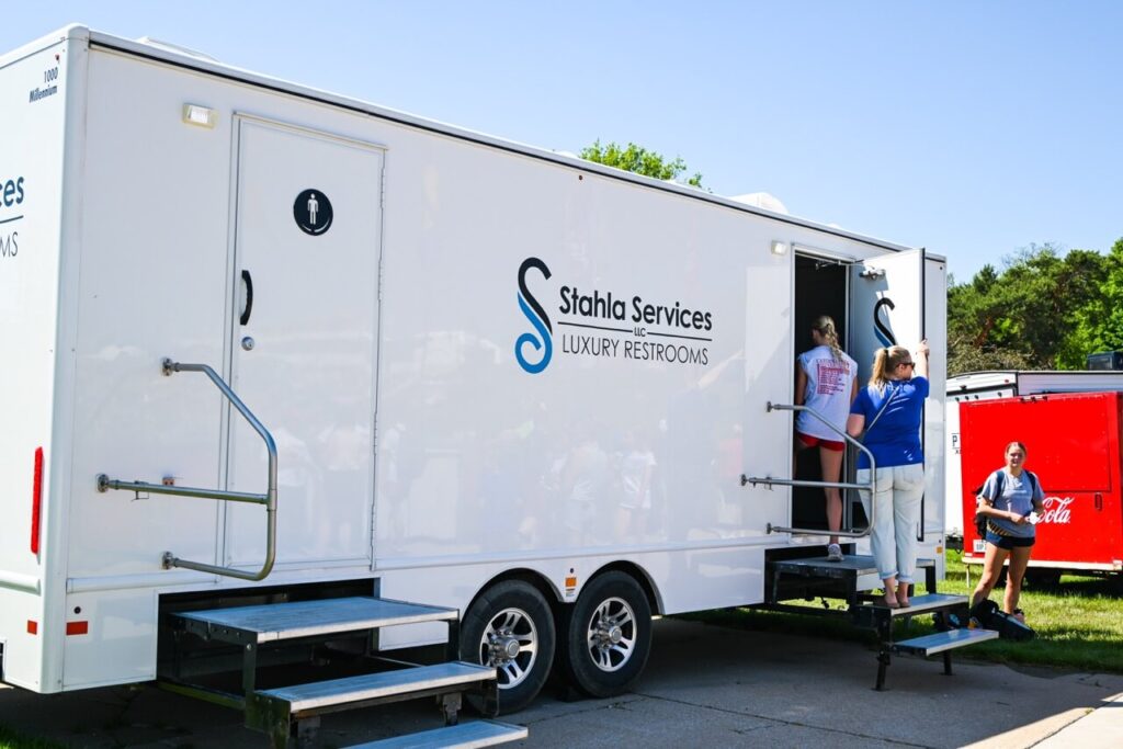 at the nsaa state track event 2024, a large white trailer labeled "stahla services luxury restrooms" is set up outdoors. two women are entering through separate doors while another woman stands nearby, holding the door open, enhancing the attendee experience with top notch restroom trailers.