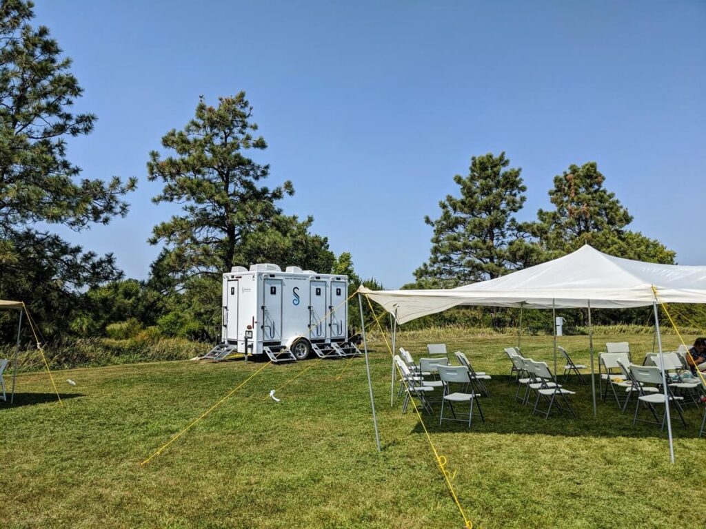 portable restrooms, including 4 stall restroom trailer rentals, and arranged folding chairs under tents set up on a grassy field with trees in the background.
