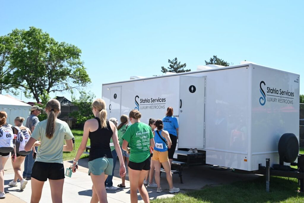a group of people in athletic wear wait in line outside a mobile restroom trailer labeled "sara's services luxury restrooms" on a sunny day, enhancing the attendee experience at the nsaa state track event 2024.