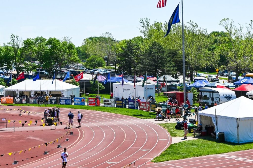 Image 3: an outdoor track meet at the nsaa state track event features runners on the curved track, spectators, tents, and flags alongside. trees and parked vehicles are visible in the background under a clear sky, enhancing the attendee experience with convenient restroom trailers on site.