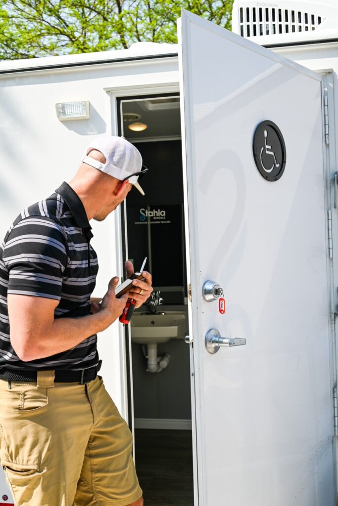 a person wearing a striped shirt and a cap is inspecting a white portable restroom marked with a wheelchair accessible symbol, holding tools. the scene has the charm of the orange city tulip festival, blending comfort and tradition with essential amenities.